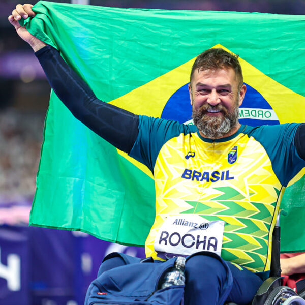 André Rocha, atleta paralímpico em cadeira de rodas, comemora os resultados do Brasil com a bandeira Nacional. Ele veste uniforme nas cores verde e amarelo nos Jogos de Paris 2024.