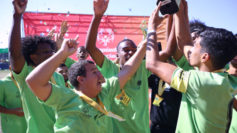 Atletas da delegação brasileira comemoram a vitória, levantando um troféu. A equipe, que irá aos Jogos Latino-Americanos das Olimpíadas Especiais 2024, veste camisetas verdes e medalhas douradas.