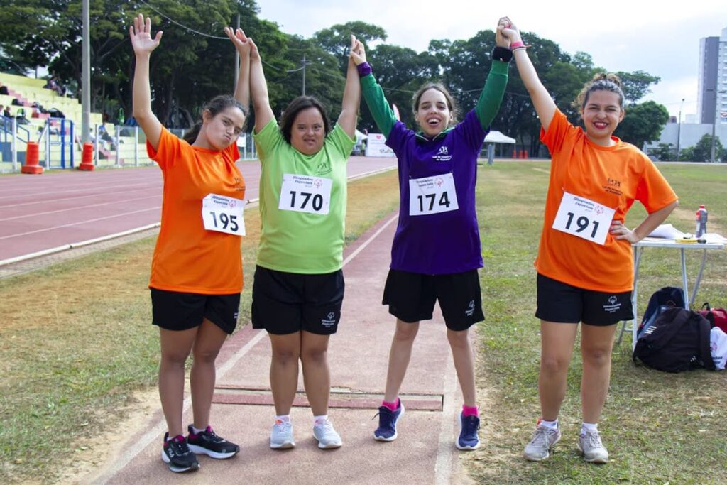 Quatro atletas brasileiras que irão aos Jogos Latino-Americanos das Olimpíadas Especiais 2024 sorriem e levantam os braços. Elas vestem camisetas de diferentes cores e números de competição.