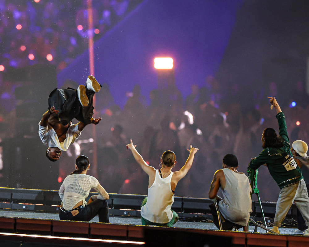 Dançarino faz acrobacia durante a festa inclusiva paralímpica de encerramento dos Jogos de Paris 2024, enquanto outros artistas observam.