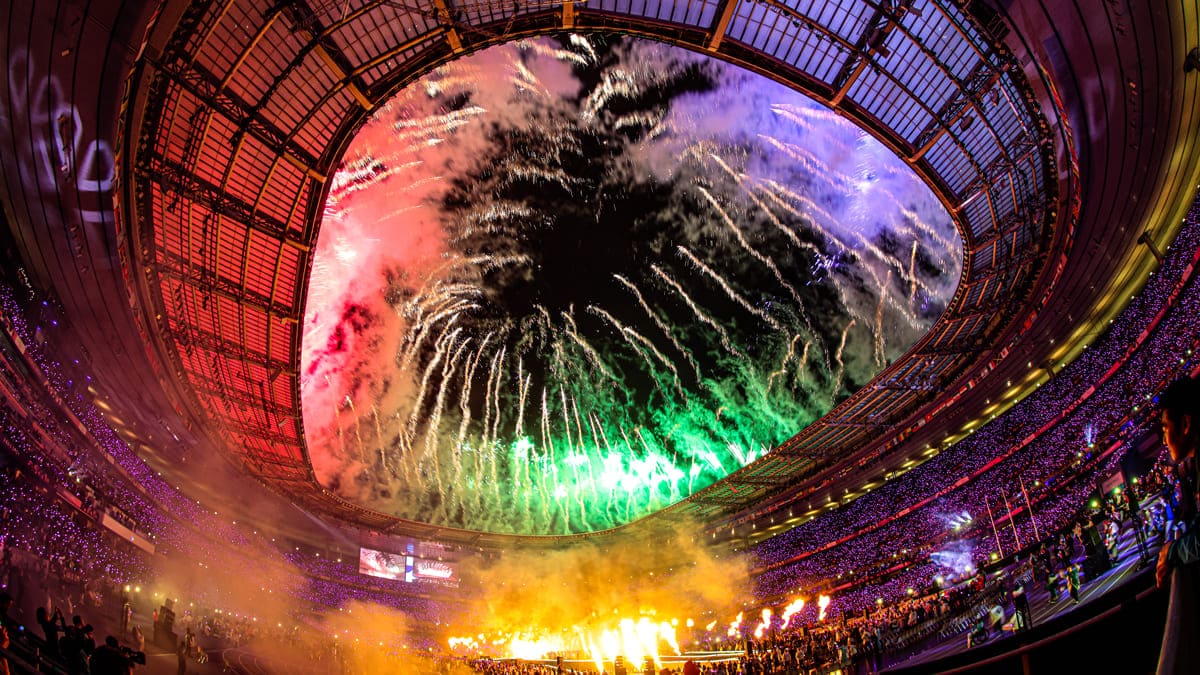 Fogos de artifício coloridos explodem no céu sobre o estádio durante a cerimônia de encerramento dos Jogos Paralímpicos de 2024, enquanto o público assiste em meio a luzes e efeitos de fumaça.