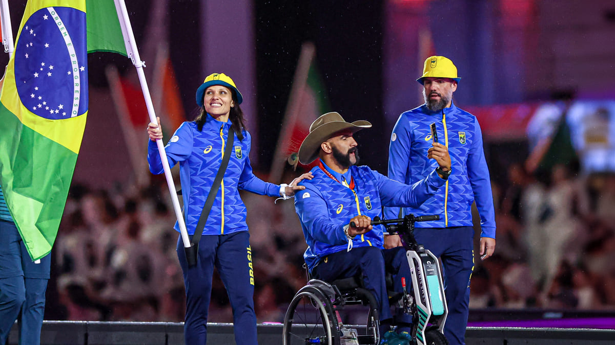 Carol Santiago e Fernando Rufino na cerimônia de encerramento dos Jogos Paralímpicos de Paris 2024, consagrando o Brasil como potência paralímpica. Carol segura a bandeira brasileira e sorri, enquanto Fernando está em cadeira de rodas usando um chapéu de cowboy.