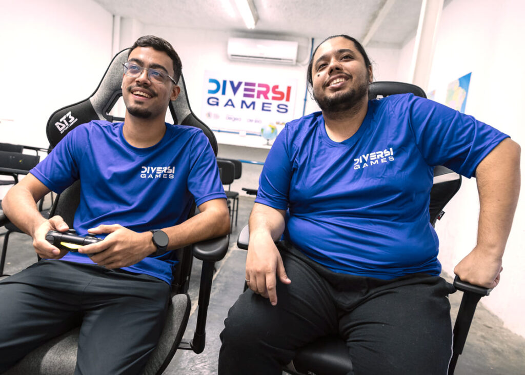 Arthur Lopes e Gabriel Nogueira, atletas de eSports, jogam em cadeiras gamer, ambos usando camisetas azuis com o logo da DiversiGames.