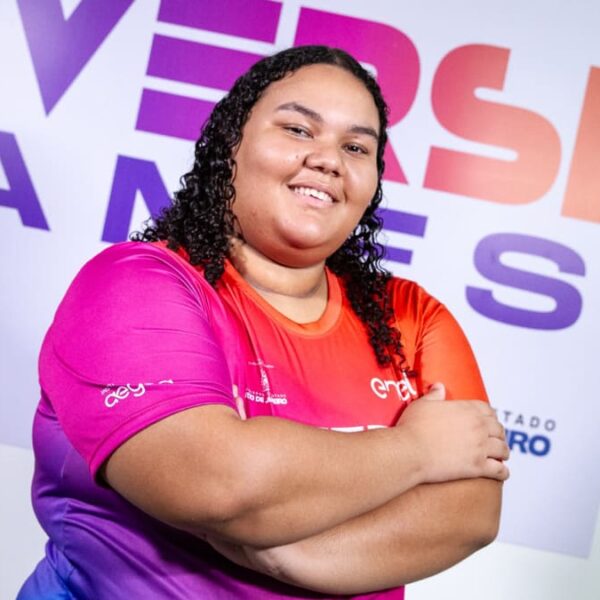 A atleta de eSports Maria Luiza dos Santos Souza, a Maluzzita, sorrindo e de braços cruzados em frente a um banner da DiversiGames.