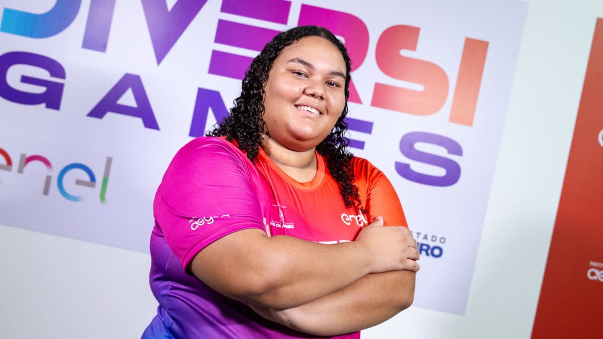 A atleta de eSports Maria Luiza dos Santos Souza, a Maluzzita, sorrindo e de braços cruzados em frente a um banner da DiversiGames.