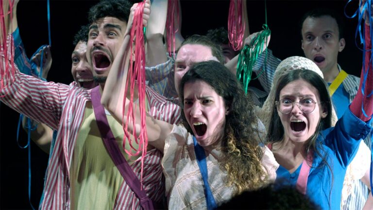 Integrantes da Orquestra Brasileira de Cantores Cegos em cena teatral, expressando emoção com fitas coloridas nas mãos.