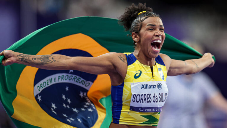 Rayane Soares da Silva comemora emocionada com a bandeira do Brasil após vitória, vestindo uniforme amarelo e azul, garantindo o país no top 5 Paralímpico.