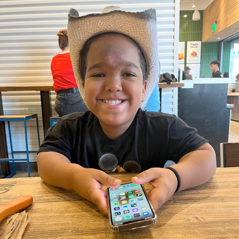 Bernard, criança de 11 anos com acondroplasia, sorri enquanto segura um celular em um restaurante. Ele está usando um chapéu de cowboy e uma camiseta preta.