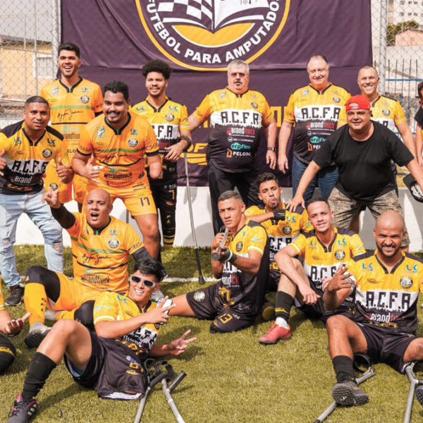 A equipe do Acre Clube de futebol de amputados, favorita da Série B do Brasileirão, posando para foto em um campo, com uniformes amarelos e pretos. Alguns estão sentados e sorrindo, segurando muletas.