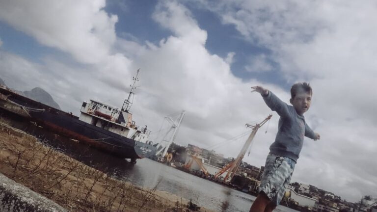 Criança brinca com os braços abertos em frente a um navio no Parque da Prainha, Vila Velha. Foto por Jarlison Gardiman, da nova exposição da Escola de Fotógrafos Cegos.