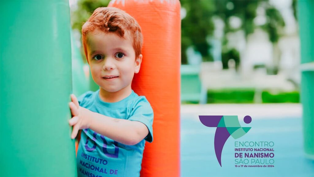 Foto de um menino com nanismo sorrindo e segurando uma estrutura inflável colorida, usando uma camiseta azul. No canto inferior direito, o logotipo da 7ª edição do Encontro Instituto Nacional de Nanismo em São Paulo, de 15 a 17 de novembro de 2024.