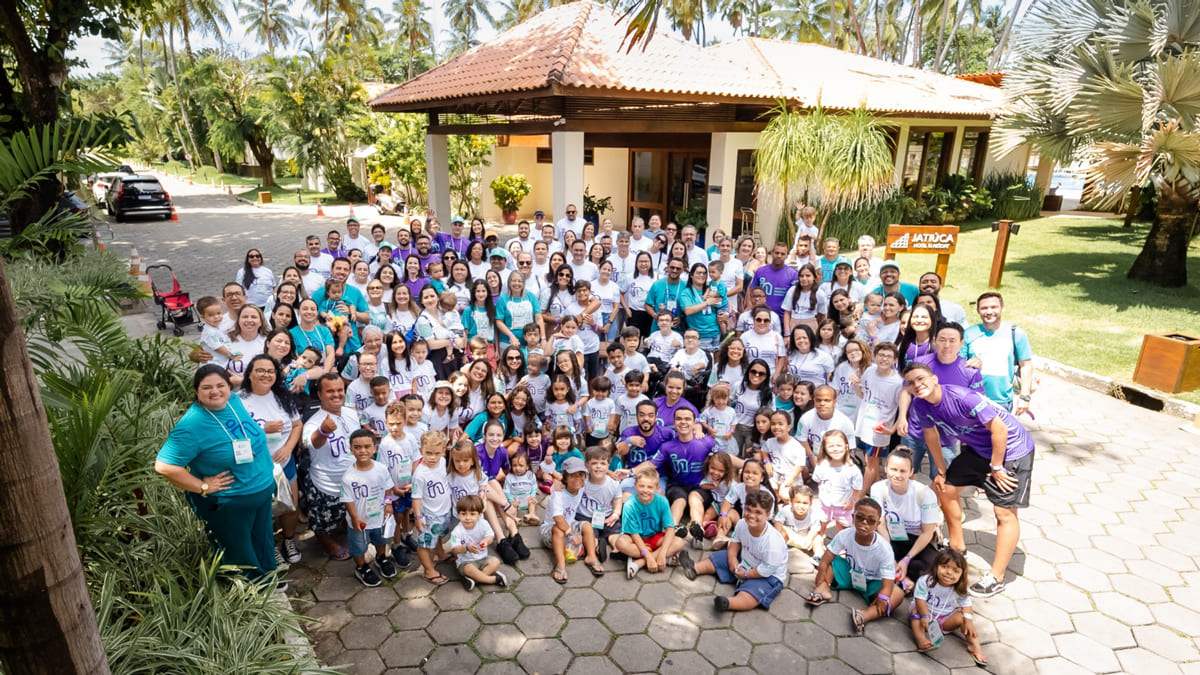 Grupo da edição anterior do Encontro sobre Nanismo, realizada no Nordeste, mostrando dezenas de participantes de várias idades posando juntos em frente a um prédio, cercados por uma paisagem tropical com palmeiras.