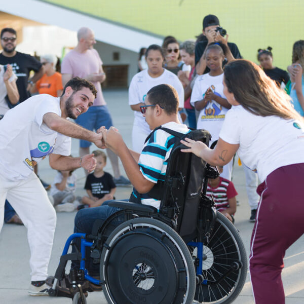 Breno, um estudante em cadeira de rodas, participa de uma roda de capoeira como parte de um projeto educacional alternativo, cercado por outras pessoas participantes.