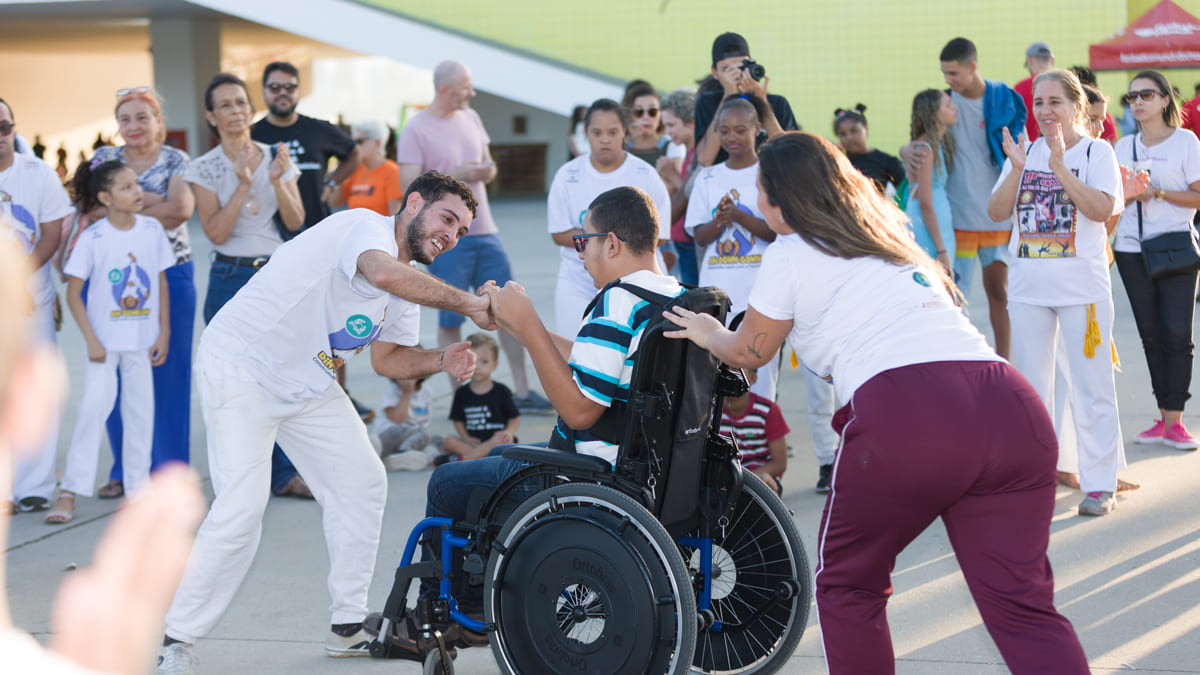 Breno, um estudante em cadeira de rodas, participa de uma roda de capoeira como parte de um projeto educacional alternativo, cercado por outras pessoas participantes.