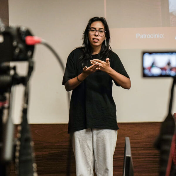 Mulher de óculos e roupas casuais sinaliza em Libras enquanto fala em frente a uma câmera. Ao fundo, é possível ver um projetor e equipamentos de filmagem desfocados.