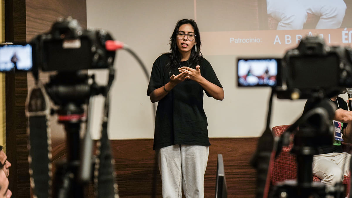 Mulher de óculos e roupas casuais sinaliza em Libras enquanto fala em frente a uma câmera. Ao fundo, é possível ver um projetor e equipamentos de filmagem desfocados.