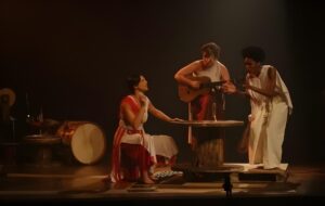 Cena da peça "Hereditária", com três atrizes no palco representando as Moiras, uma delas tocando violão enquanto as outras interagem ao redor de uma mesa de madeira.