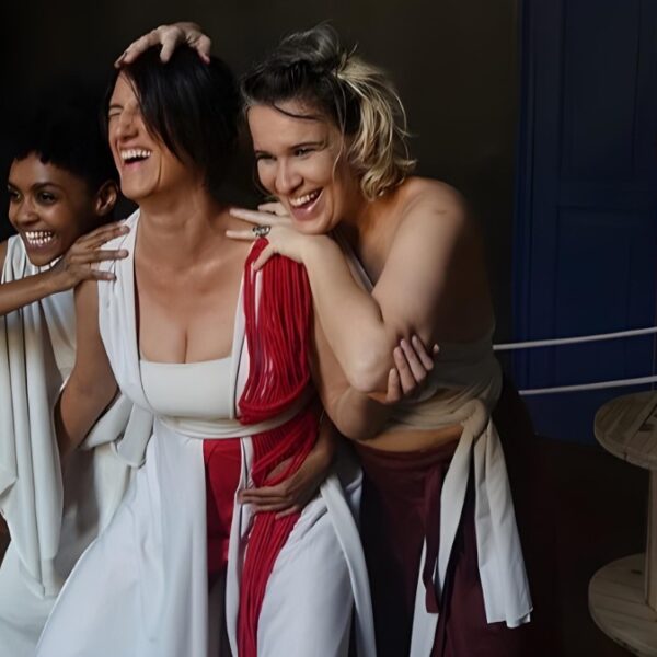Três atrizes caracterizadas como Moiras da mitologia grega riem juntas em cena da peça Hereditária. Elas vestem figurinos em branco e vermelho, e ao fundo há um grande carretel de madeira com fios estendidos.