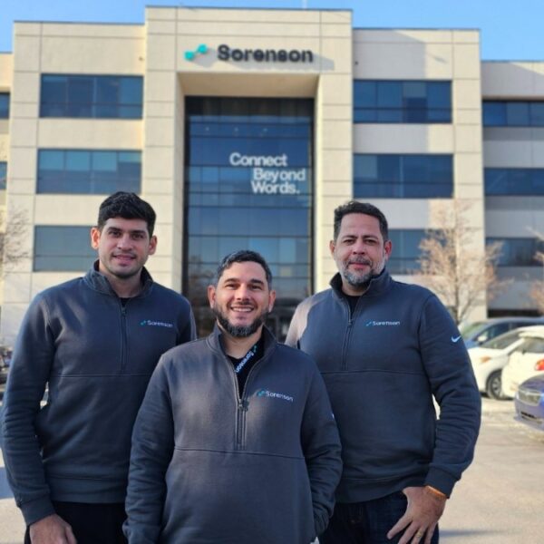 Fundadores da Hand Talk posam em frente ao prédio da Sorenson. Eles vestem jaquetas com o logotipo da empresa, e ao fundo há carros estacionados e árvores sem folhas.
