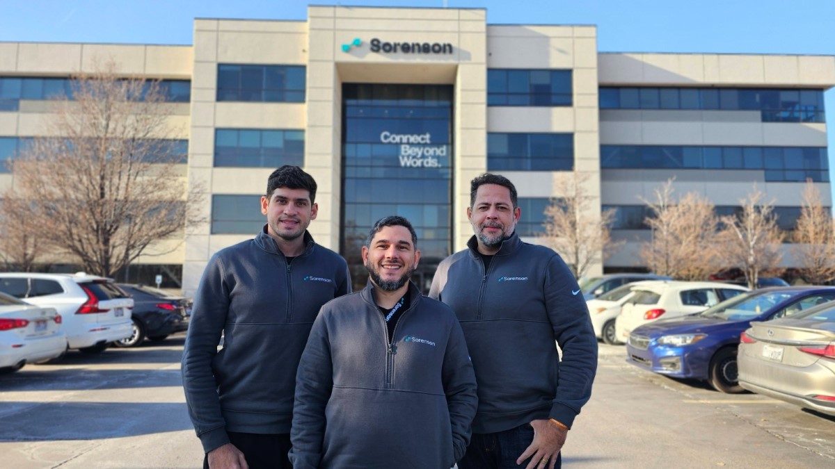 Fundadores da Hand Talk posam em frente ao prédio da Sorenson. Eles vestem jaquetas com o logotipo da empresa, e ao fundo há carros estacionados e árvores sem folhas.