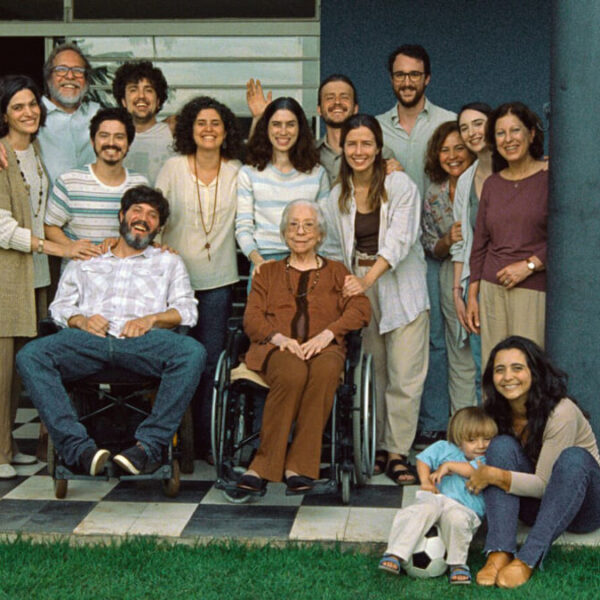Cena do filme Ainda Estou Aqui, com um grupo de pessoas reunidas na varanda de uma casa, com uma pessoa em cadeiras de rodas no centro e uma mulher abraçando uma criança no chão.