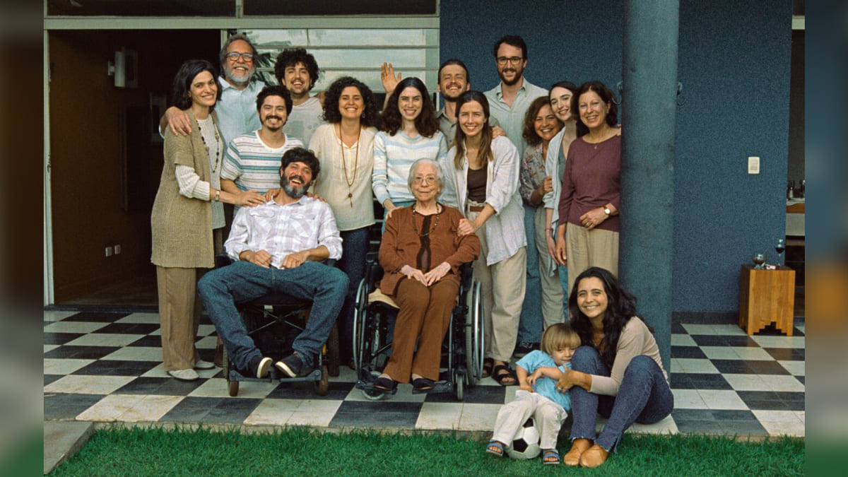 Cena do filme Ainda Estou Aqui, com um grupo de pessoas reunidas na varanda de uma casa, com uma pessoa em cadeiras de rodas no centro e uma mulher abraçando uma criança no chão.