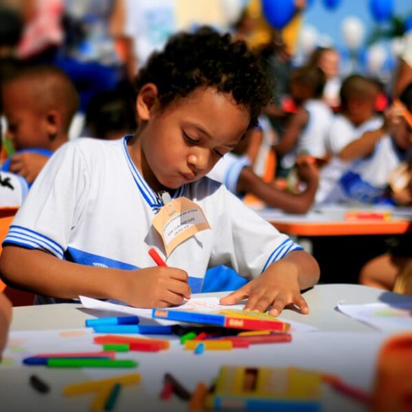 Criança negra com uniforme escolar branco e azul desenha com lápis de cor em uma mesa ao ar livre, cercada por outros alunos. Logo do Instituto Cultiva.