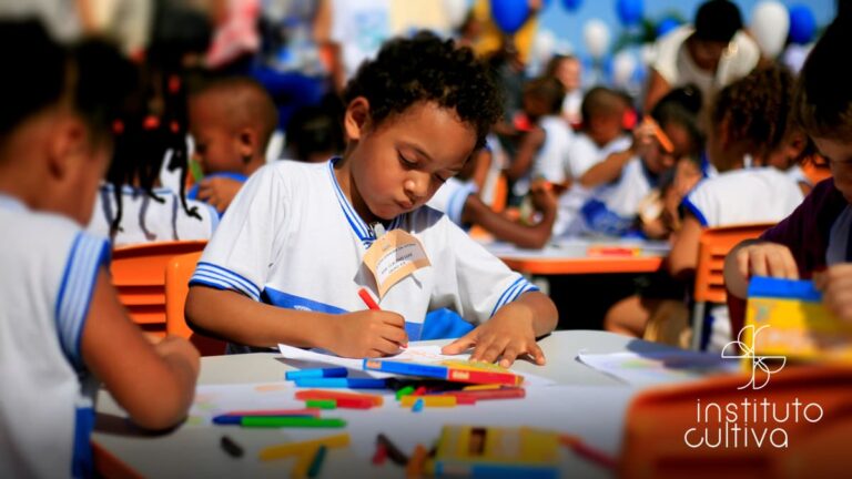 Criança negra com uniforme escolar branco e azul desenha com lápis de cor em uma mesa ao ar livre, cercada por outros alunos. Logo do Instituto Cultiva.