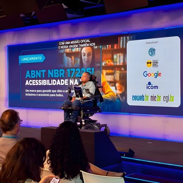 Marco Pellegrini, em cadeira de rodas, fala no lançamento da norma ABNT que eleva o padrão da web, realizado na sede do Google em São Paulo. Ao lado, um intérprete de Libras traduz o discurso para a plateia.