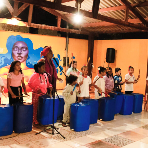Crianças da periferia e comunidade indígena tocam tambores azuis em uma oficina de percussão, acompanhadas por uma mulher com turbante vermelho e uma instrutora.