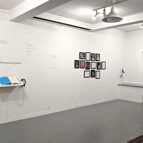 Sala de galeria com paredes brancas exibe textos tipográficos na parede, um livro azul aberto com fones de ouvido, fotos emolduradas no centro e a imagem digital de um rosto com correntes douradas à direita.