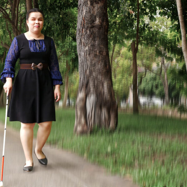 Mulher cega caminha com bengala branca em calçada arborizada, vestindo vestido preto com mangas de renda azul - Desafio de Inovação Bengalas Inteligentes.