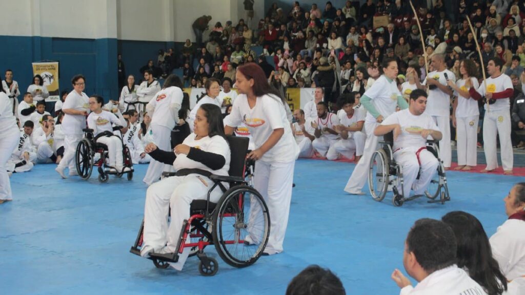 Pessoas com deficiência participam de roda de capoeira em cadeiras de rodas durante Festival Olga Kos de Inclusão Esportiva, com grande público ao fundo.