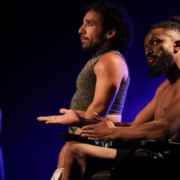 Cena da peça “Meu Corpo Está Aqui” com dois atores no palco, um deles em cadeira de rodas, sob iluminação azul. (Créditos: Fabio Alcover / Filo)