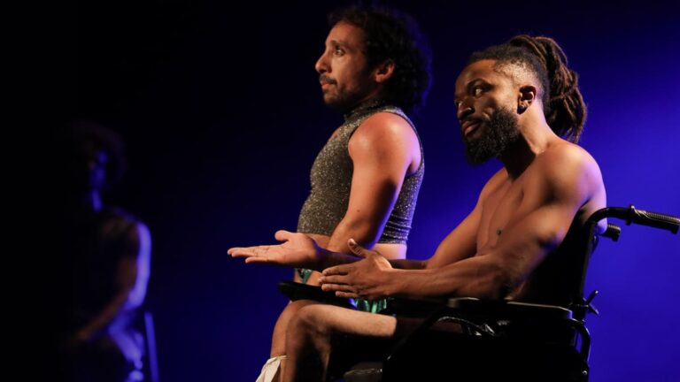 Cena da peça “Meu Corpo Está Aqui” com dois atores no palco, um deles em cadeira de rodas, sob iluminação azul. (Créditos: Fabio Alcover / Filo)