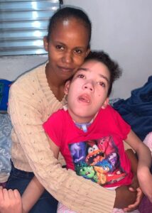 Mãe carinhosa abraça a filha com deficiência, que veste camiseta rosa com personagens coloridos e usa traqueostomia.