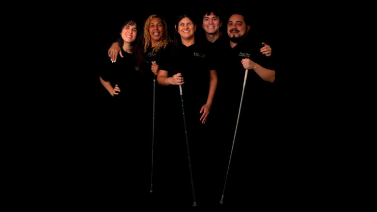 Cinco pessoas que integram o grupo Teatro Cego, três delas segurando bengalas, posam sorrindo em fundo preto. (Foto: Iasmin Dib)