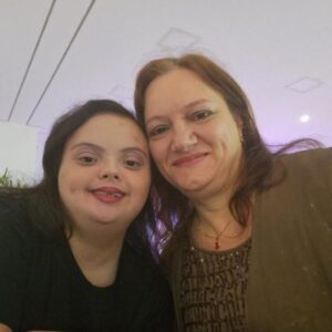 Ana e Fabiana sorrindo lado a lado em ambiente fechado com iluminação suave.