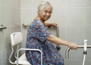 Idosa sorridente sentada em cadeira adaptada para banho, segurando barras de apoio no banheiro.
