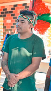 Kacique Samuel da Silva (Kacique Gog), da comunidade Kaingang, com cocar de penas vermelhas e verdes, camiseta verde e tatuagens nos braços, em ambiente comunitário.