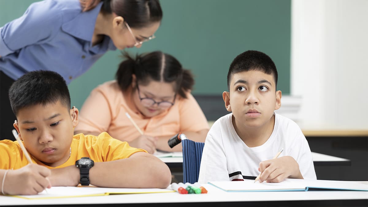 Crianças com deficiência em sala de aula, sentadas em carteiras, participando de uma atividade de escrita com apoio de uma professora ao fundo, transmitindo um ambiente de inclusão escolar. (Créditos: Depositphotos)