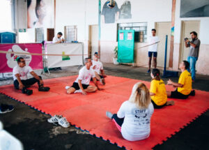 Um grupo de pessoas, algumas sentadas no chão em um tatame vermelho, participando da atividade esportiva de vôlei sentado. Ao fundo, outras pessoas observam e um homem fotografa.