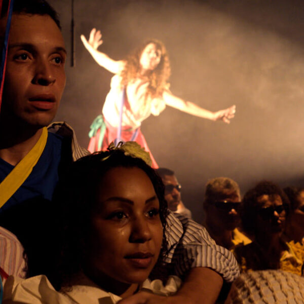 Grupo de artistas, incluindo a Orquestra Brasileira de Cantores Cegos, em cena teatral; uma mulher ao fundo, em destaque sob um facho de luz, estende os braços em movimento expressivo. (Créditos: Julio Gabriel & Cia Poéticas da Cena Contemporânea)