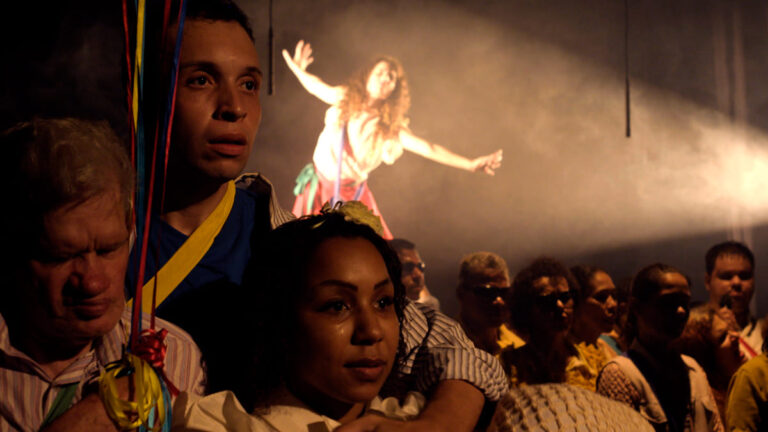 Grupo de artistas, incluindo a Orquestra Brasileira de Cantores Cegos, em cena teatral; uma mulher ao fundo, em destaque sob um facho de luz, estende os braços em movimento expressivo. (Créditos: Julio Gabriel & Cia Poéticas da Cena Contemporânea)