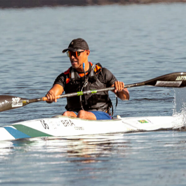 Paratleta paranaense Raphael dos Santos em sua canoa, remando em águas calmas, usando colete salva-vidas laranja e preto e óculos de sol, com a inscrição 'BRA' na proa do barco. (Foto: Divulgação)