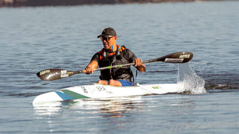 Paratleta paranaense Raphael dos Santos em sua canoa, remando em águas calmas, usando colete salva-vidas laranja e preto e óculos de sol, com a inscrição 'BRA' na proa do barco. (Foto: Divulgação)