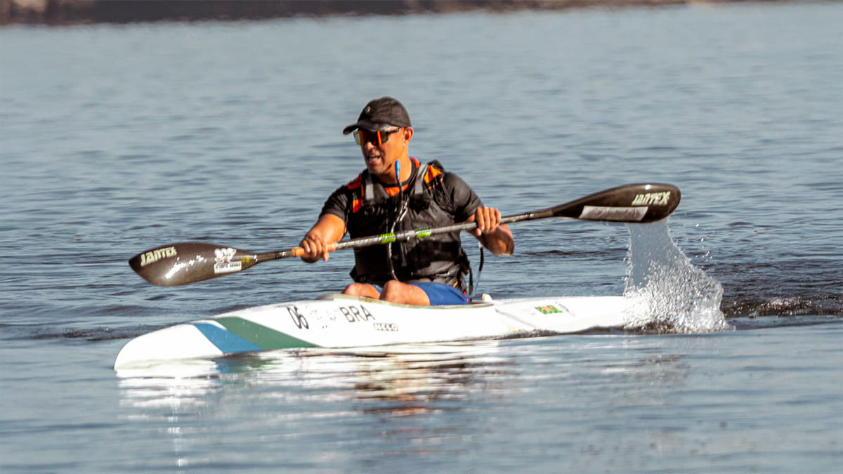 Paratleta paranaense Raphael dos Santos em sua canoa, remando em águas calmas, usando colete salva-vidas laranja e preto e óculos de sol, com a inscrição 'BRA' na proa do barco. (Foto: Divulgação)