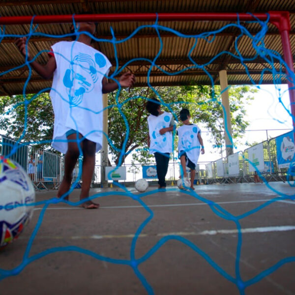 Crianças, algumas descalças e usando camisetas brancas da Virada Esportiva Inclusiva, jogam futebol em um gol com rede azul. Uma bola de futebol está em primeiro plano, dentro da rede. O ambiente é um espaço aberto com cobertura. (Foto: Ricardo Lima)