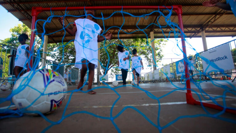 Crianças, algumas descalças e usando camisetas brancas da Virada Esportiva Inclusiva, jogam futebol em um gol com rede azul. Uma bola de futebol está em primeiro plano, dentro da rede. O ambiente é um espaço aberto com cobertura. (Foto: Ricardo Lima)