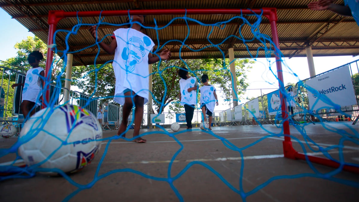 Crianças, algumas descalças e usando camisetas brancas da Virada Esportiva Inclusiva, jogam futebol em um gol com rede azul. Uma bola de futebol está em primeiro plano, dentro da rede. O ambiente é um espaço aberto com cobertura. (Foto: Ricardo Lima)